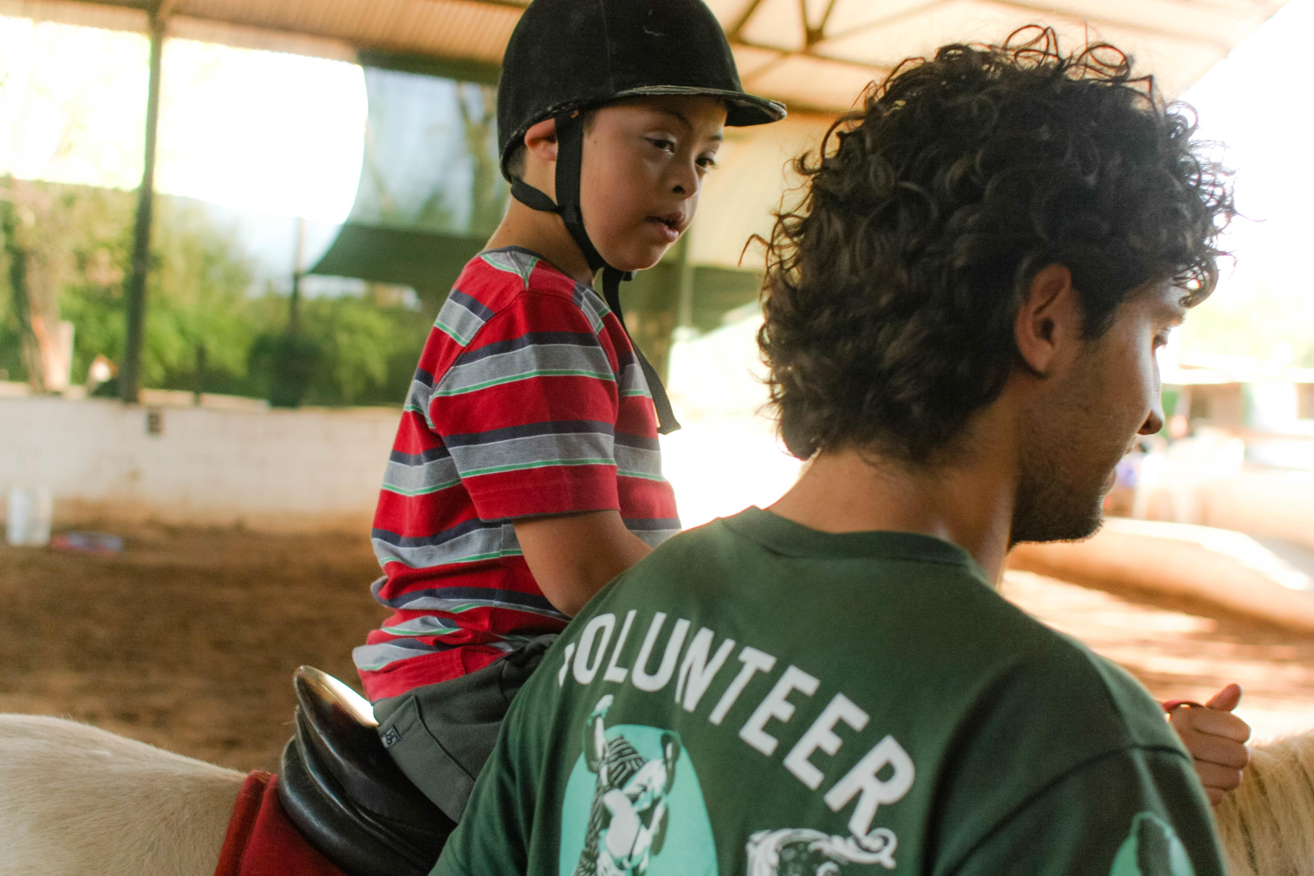 Projects Abroad Equine Therapy intern helps a young boy ride a horse in Argentina. Male Equine Therapy intern working with children in Argentina leads a horse ridden by a child during a therapy session.